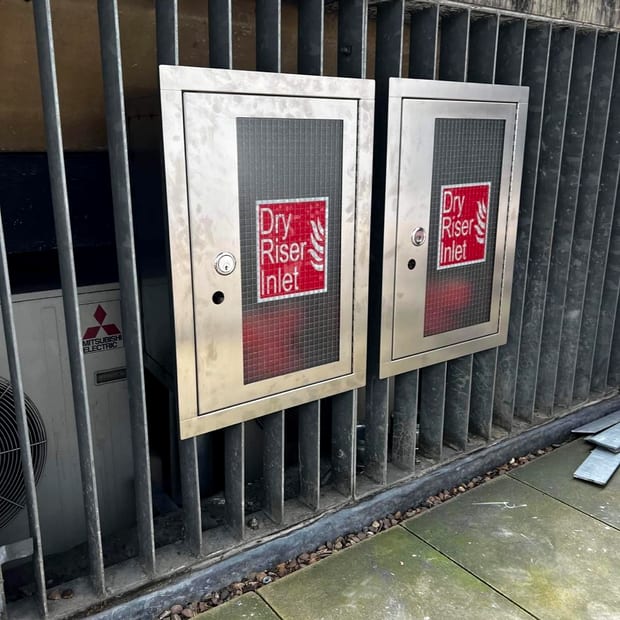 Two locked stainless steel dry riser inlet cabinets with red Dry Riser Inlet signage on a louvred plant wall