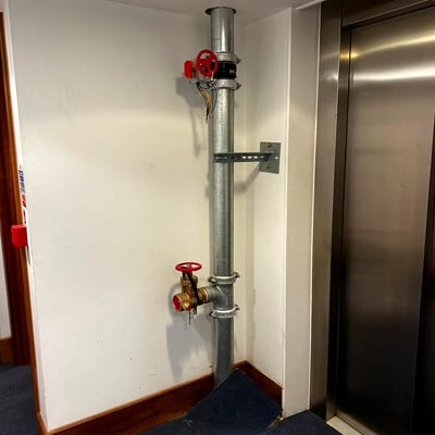 A dry riser landing valve with its red handwheel on a galvanised riser beside a lift in a residential lobby