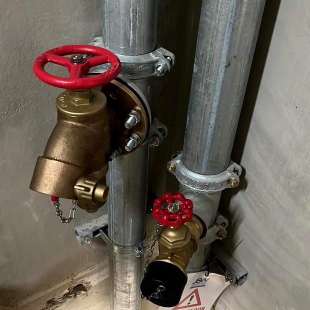Two brass wet riser landing valves with red handwheels and capped outlets on galvanised risers